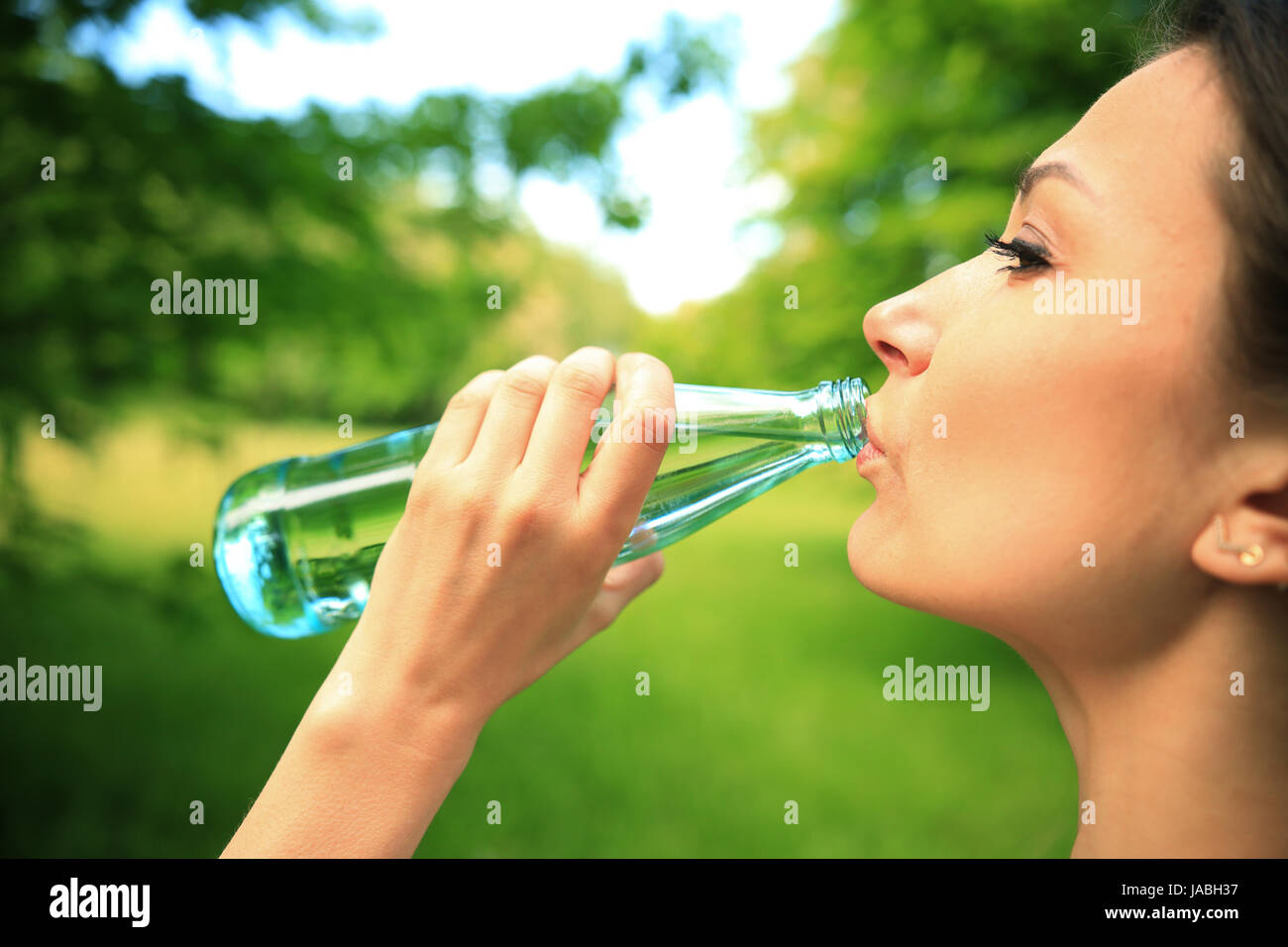 Girl drinks water close-up. Young woman with bottle of fitness water in green park. Healthy lifestyle backdrop. Stock Photo