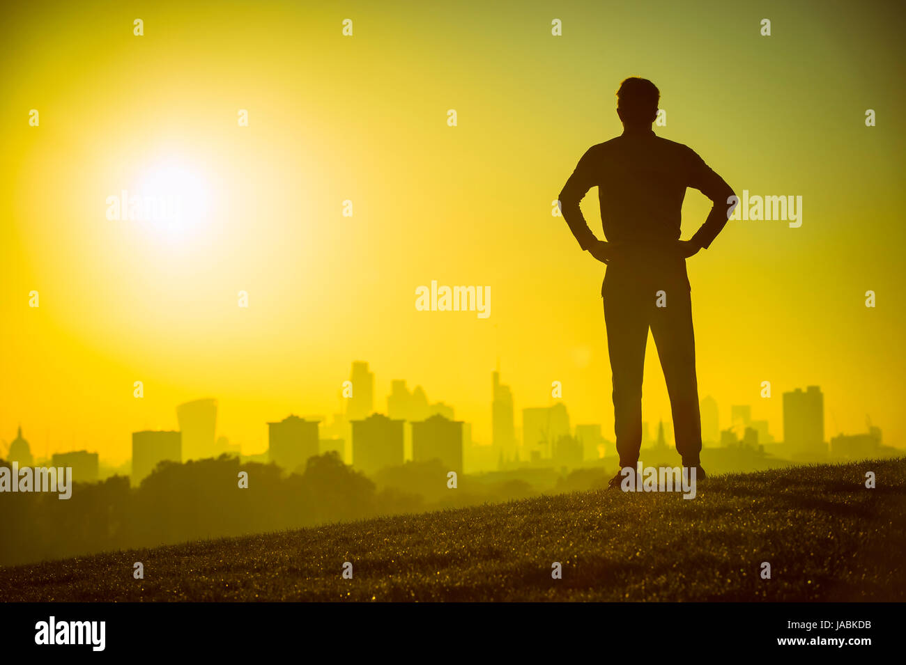 Silhouette of a defiant man with hands on hips looking out at the sunrise city skyline. Stock Photo