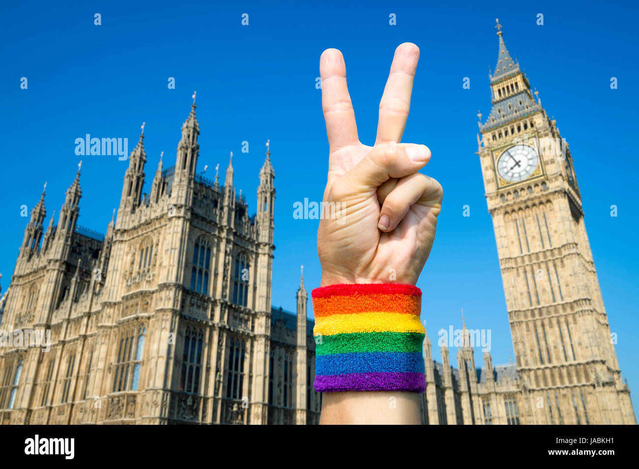Hand wearing gay pride rainbow flag wristband holding up a victory/peace sign gesture in front of the Houses of Parliament at Westminster, London, UK Stock Photo
