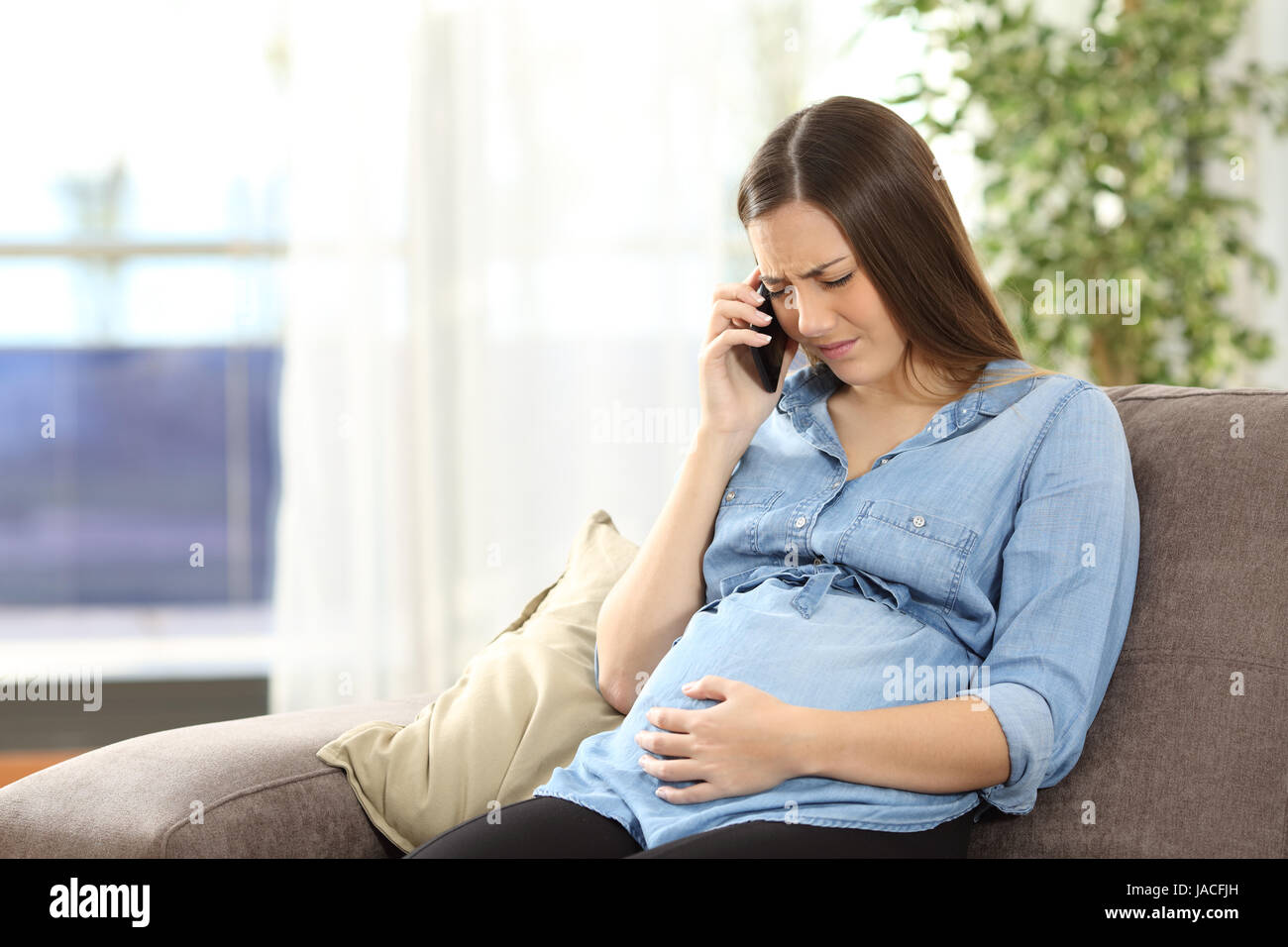 Young pregnant woman suffering belly ache and calling on the phone sitting on a couch in the living room in a house interior Stock Photo