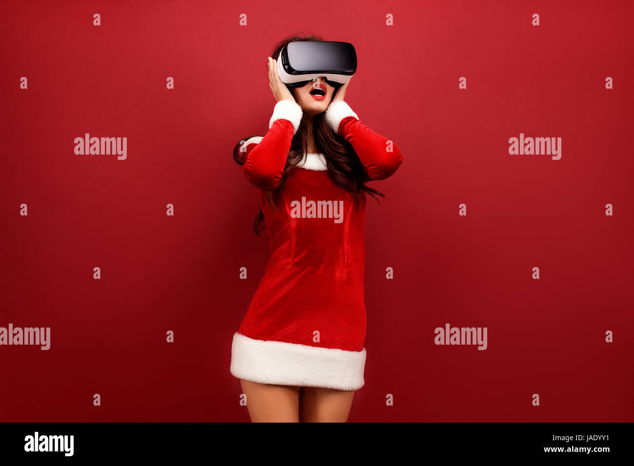 Portrait of young brunette woman in red velvet dress using virtual reality headset and touching head with arms Stock Photo