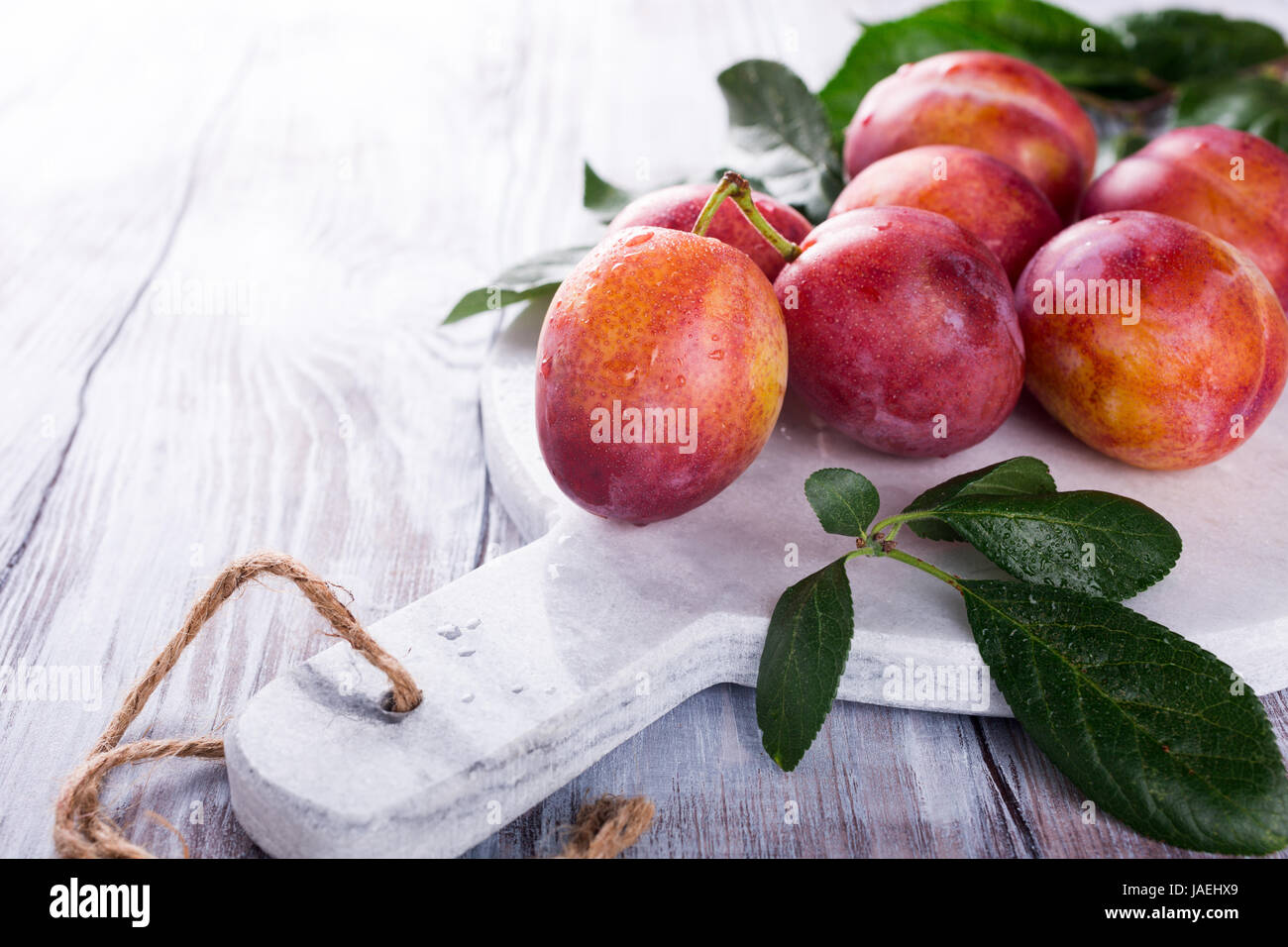 Fresh plums on marble cutting board on old white wooden background. Selective focus. Healthy food concept. Copy space. Stock Photo