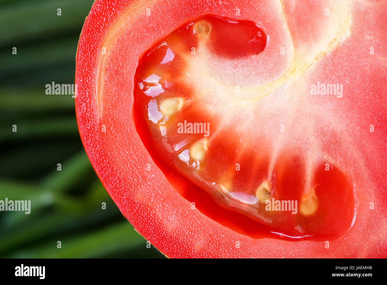 Extreme close up of a half sliced tomato against blurred scallion leaves. Abstract macro texture food background Stock Photo