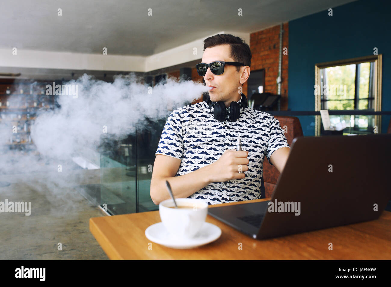Young handsome hipster man with beard sitting in cafe with a cup of coffee, vaping and releases a cloud of vaporsitting. Working at laptop and having  Stock Photo