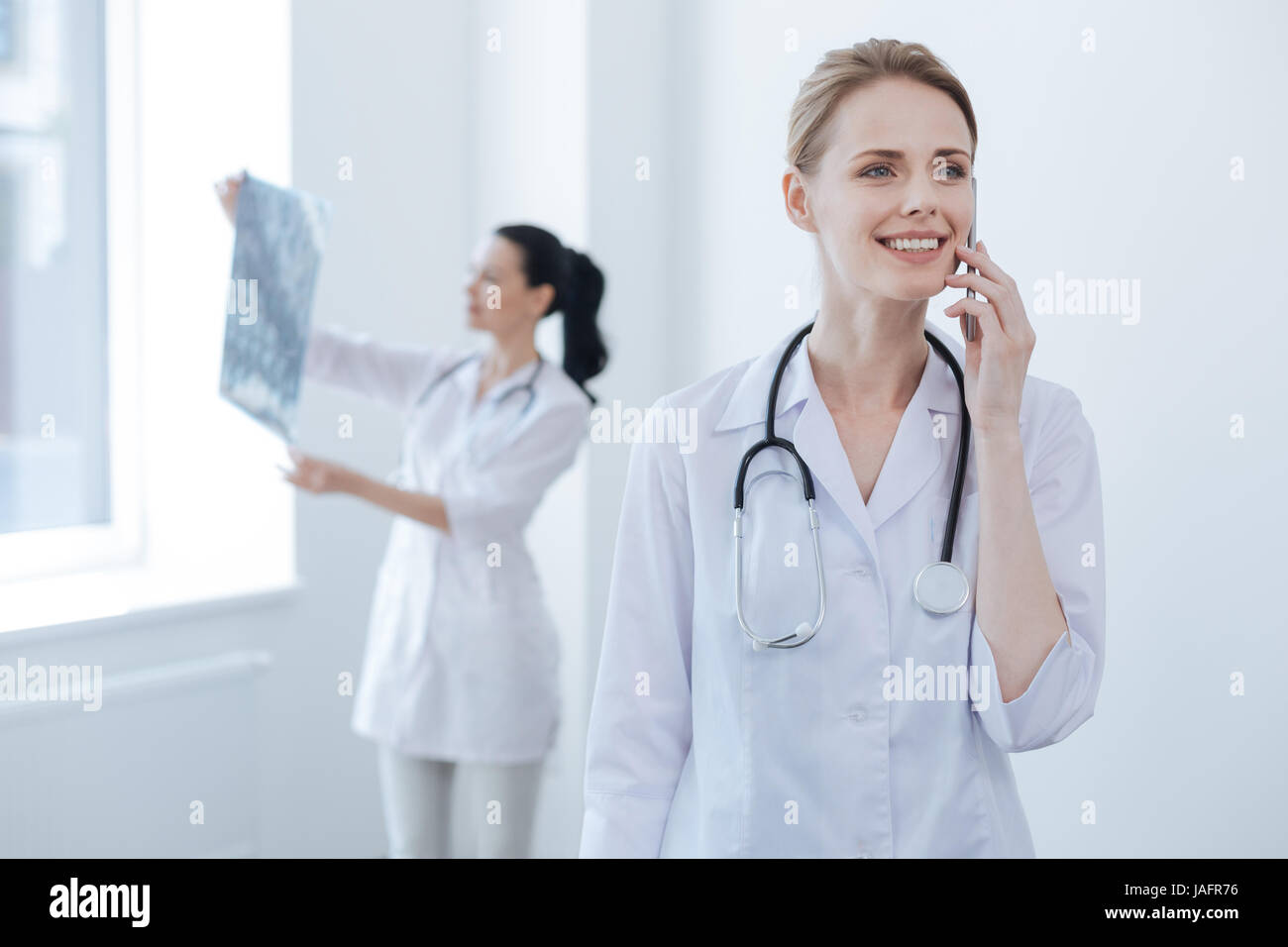 Full of joy. Proficient glad mature neurologist working at the medical lab and enjoying conversation on the smartphone while her colleague holding bra Stock Photo