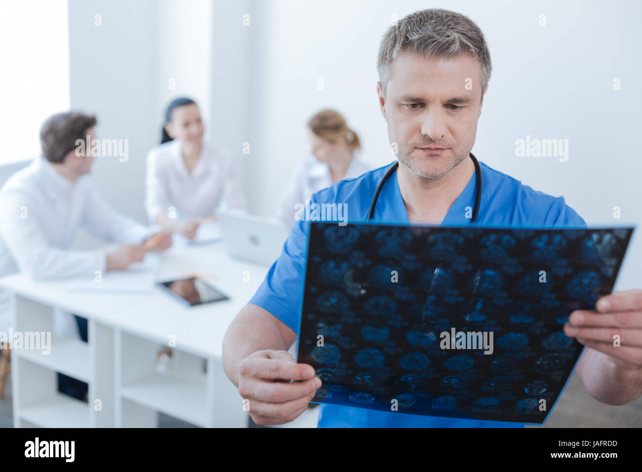Examining magnetic resonance image. Concentrated mature experienced neurologist working at the lab and examining mrt scan results while colleagues enj Stock Photo