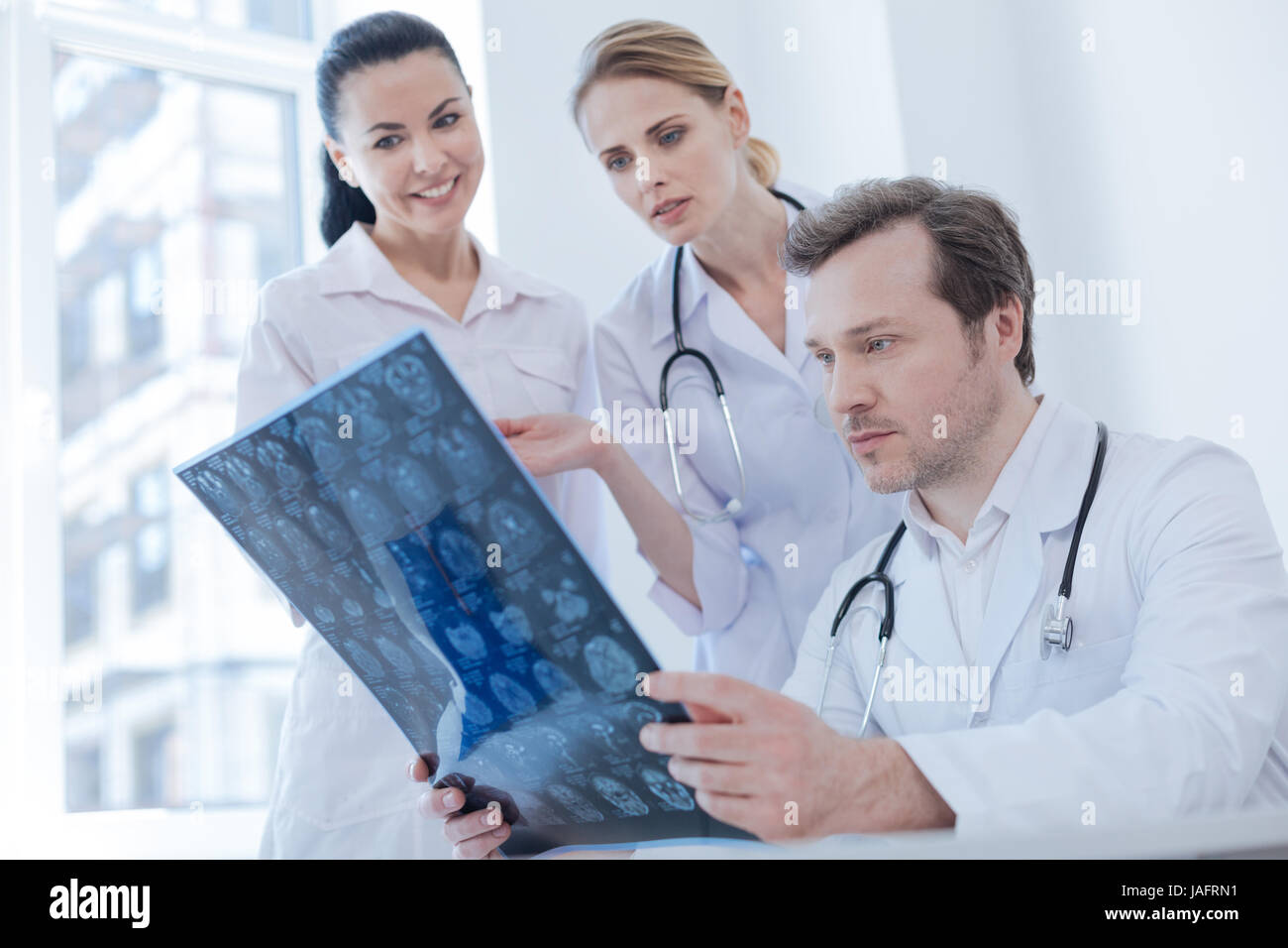 Detecting pathology . Qualified mature attentive neurologist working and analyzing brain x ray image at the laboratory while colleagues sharing opinio Stock Photo