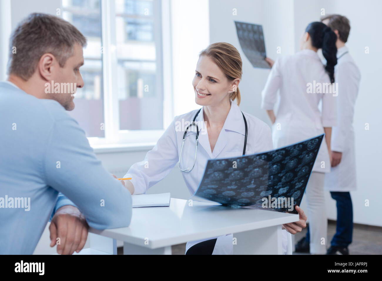 Providing checkup of your health. Confident qualified kind neurologist enjoying appointment with patient and analyzing mrt scan while colleagues enjoy Stock Photo