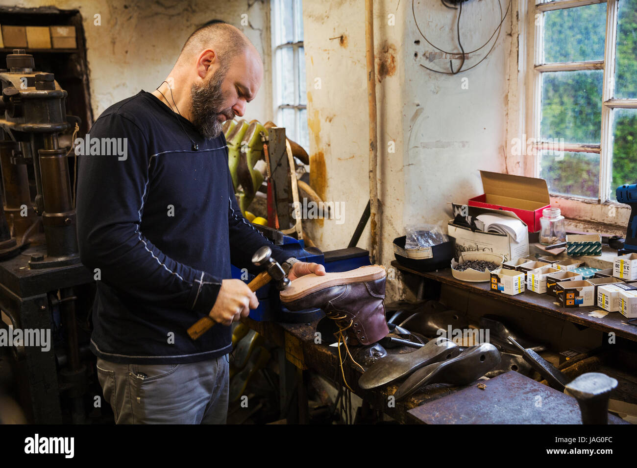 Man standing in a shoemaker's workshop by a window, using a hammer on the soles of a work boot. Stock Photo