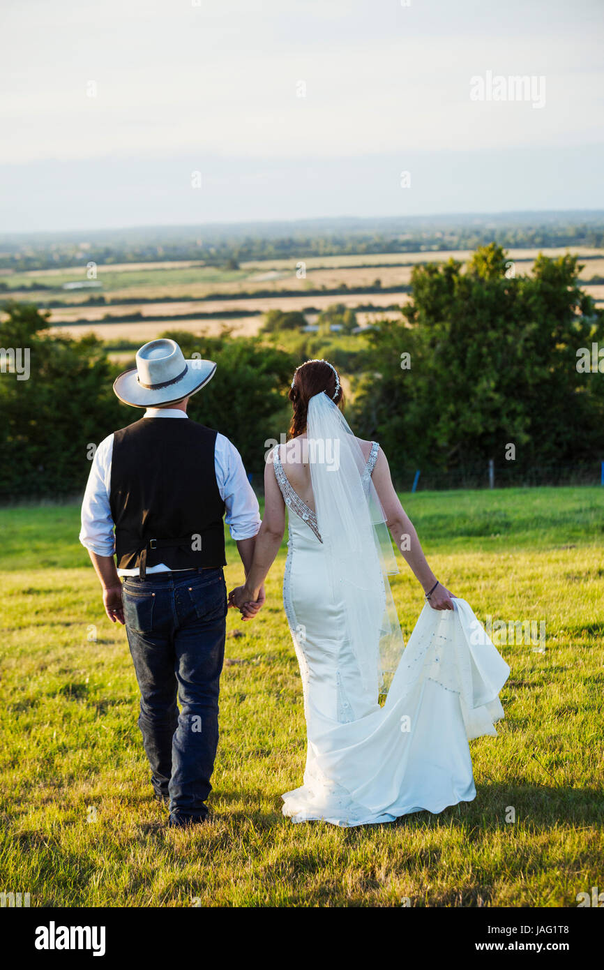 Rear view of bride and groom walking hand in hand across a grassy slope ...