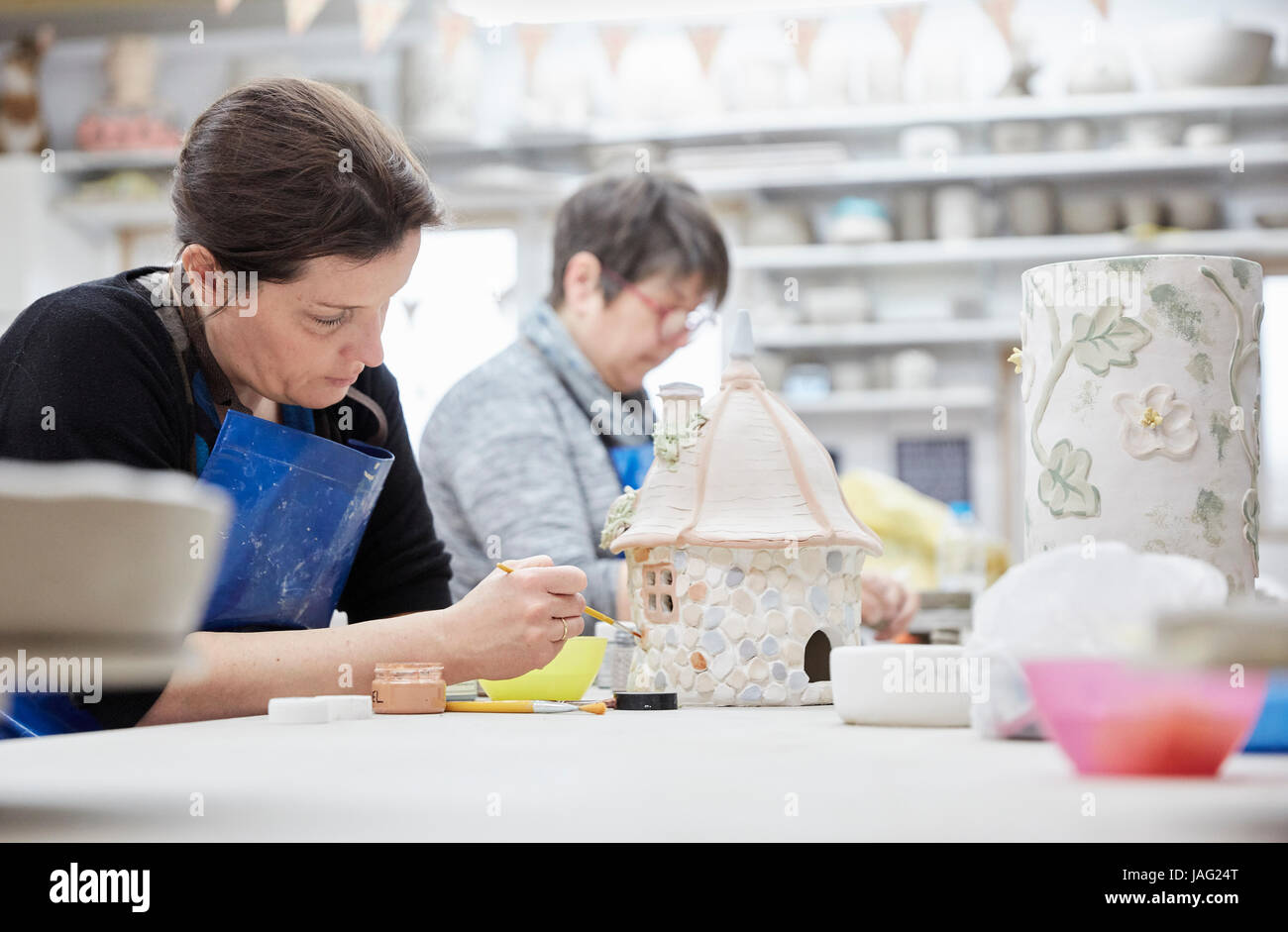 Two women seated at a workbench in a pottery studio, working on decorated clay objects. Stock Photo