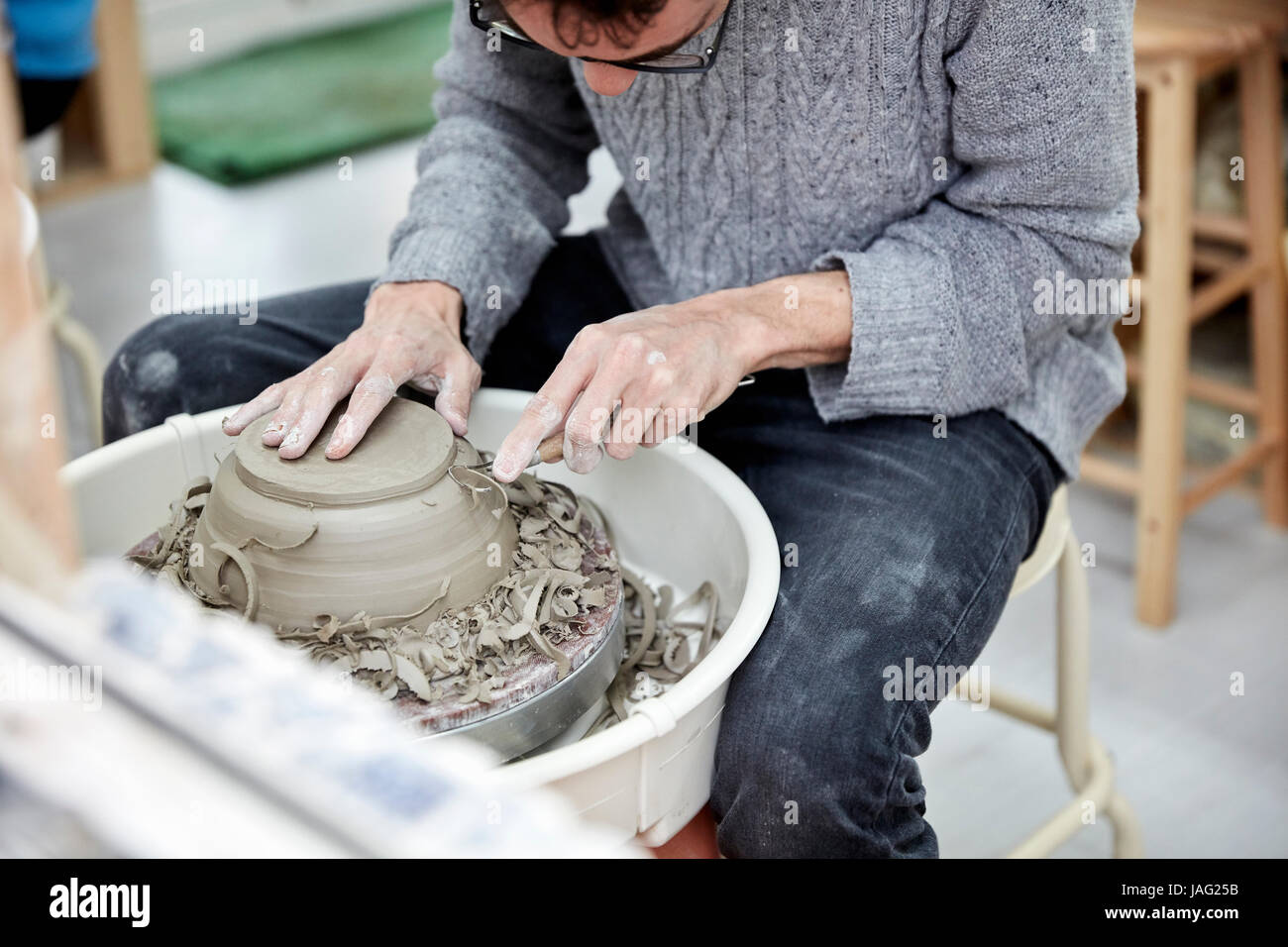 A man using a pottery wheel, shaping a pot base with a small handheld tool shaving off excess clay. Stock Photo