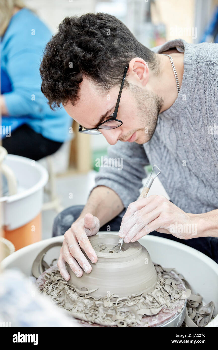 A man seated at a potter's wheel working and shaping a clay pot by removing excess clay. Stock Photo