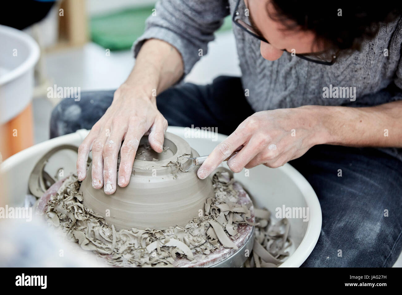 A man seated at a potter's wheel working and shaping a clay pot by removing excess clay. Stock Photo