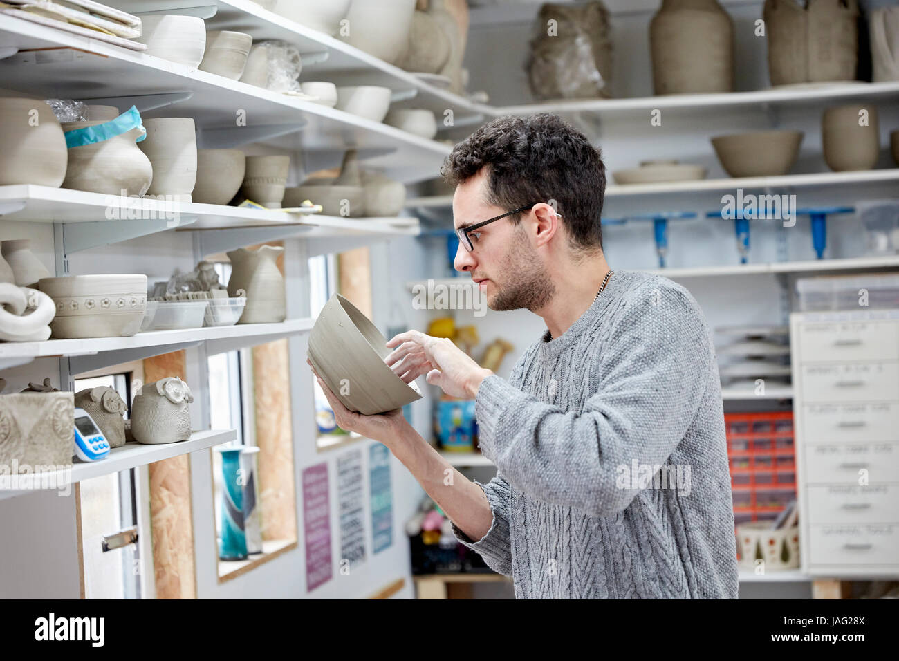 A man inspecting a clay pot, before firing. Shelves in a pottery studio full of pots, work in progress. Stock Photo