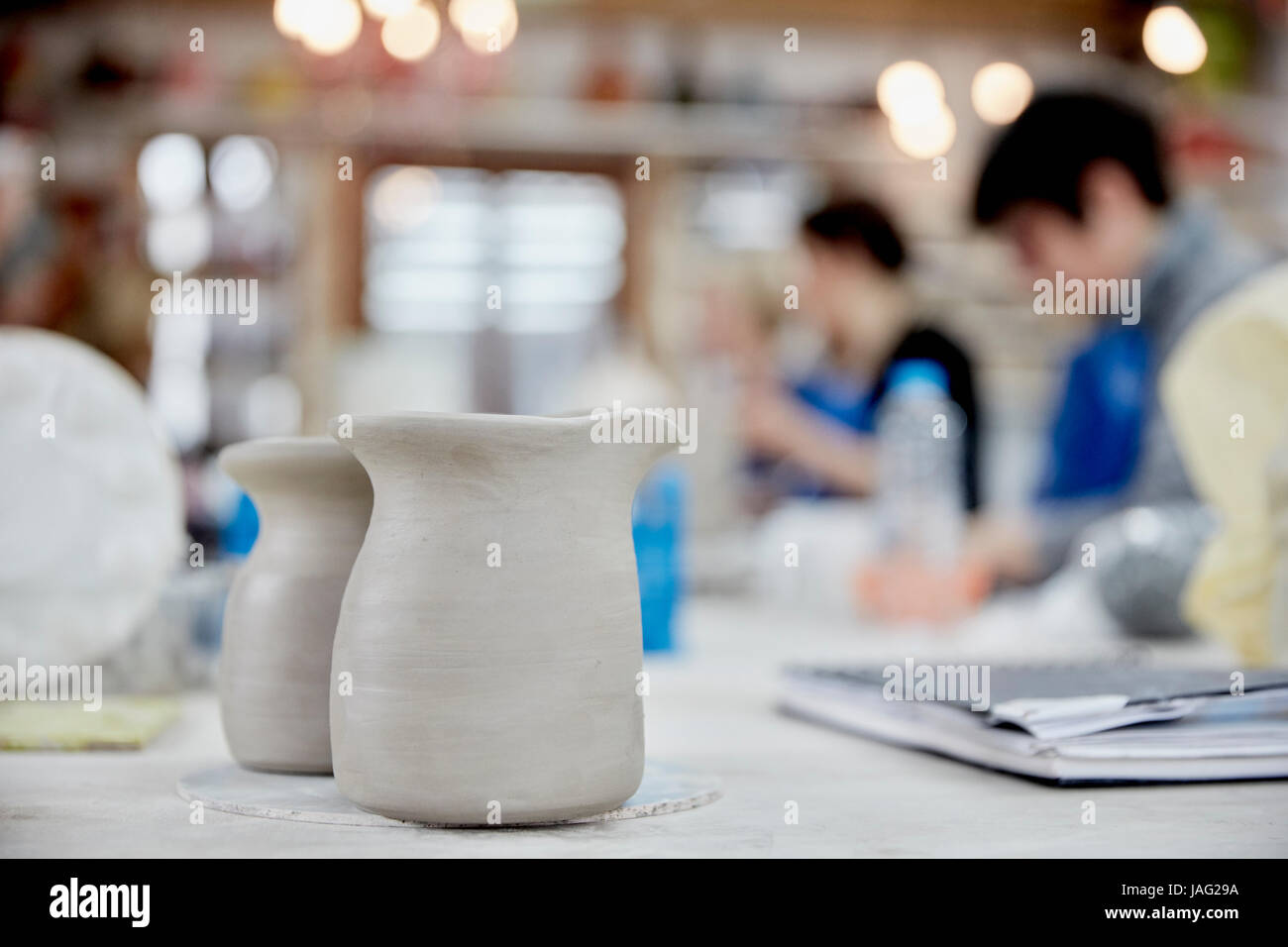 Two clay jugs in the foreground. A ceramics class taking place, people seated at a workbench in a pottery studio. Stock Photo