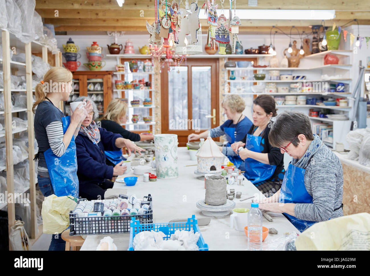 A group of people seated at a workbench in a pottery workshop, handbuilding clay objects. A woman with a cup of tea. Stock Photo