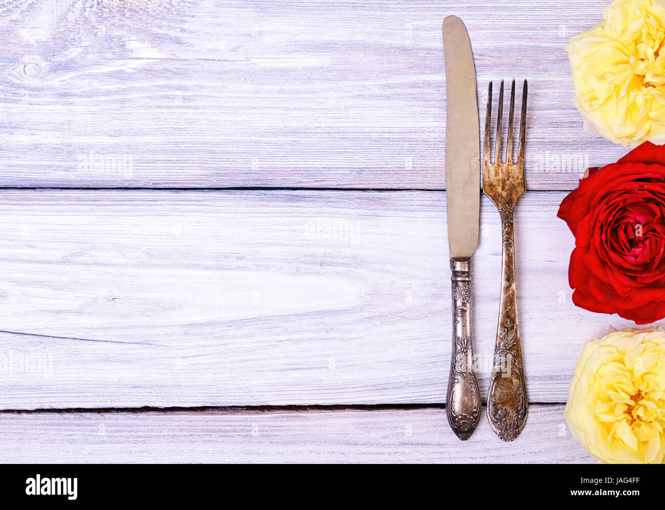 Iron cutlery fork and knife on a white wooden table, decorated with roses, empty space on the left Stock Photo