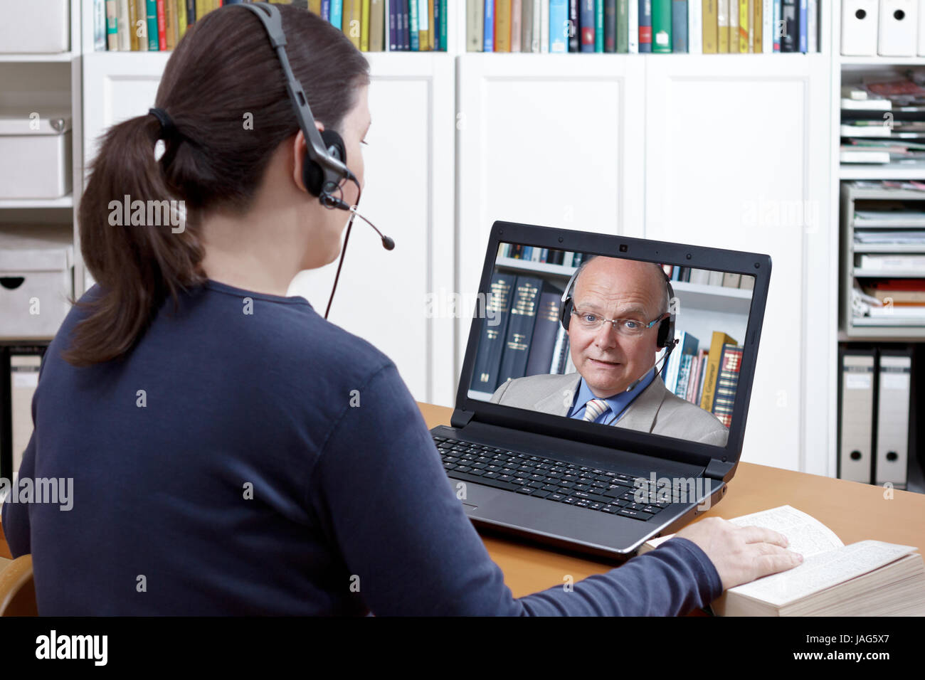 Woman with headset and book at her desk in front of her laptop having an online chat with her prof, text space Stock Photo