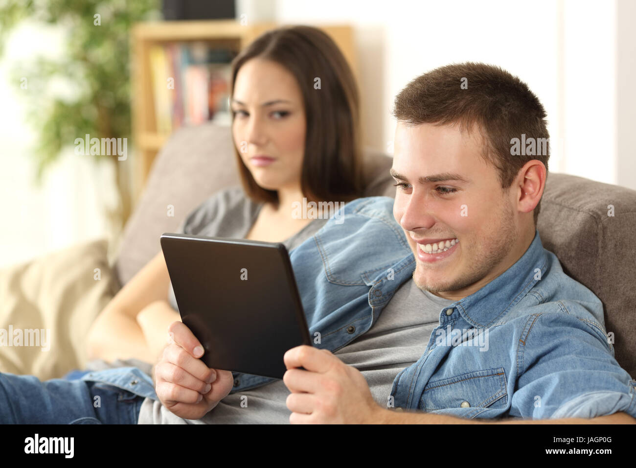 Husband watching online content in a tablet and ignoring to his angry wife who is seated beside him on a sofa at home Stock Photo