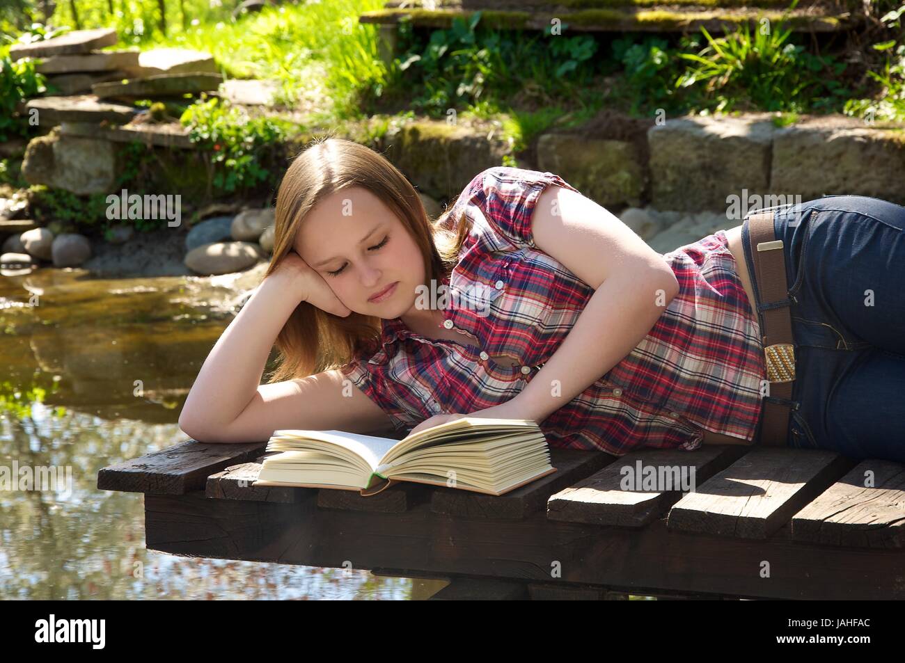 Junge Frau liest ein Buch am Gartenteich und liegt auf einem Steg Stock