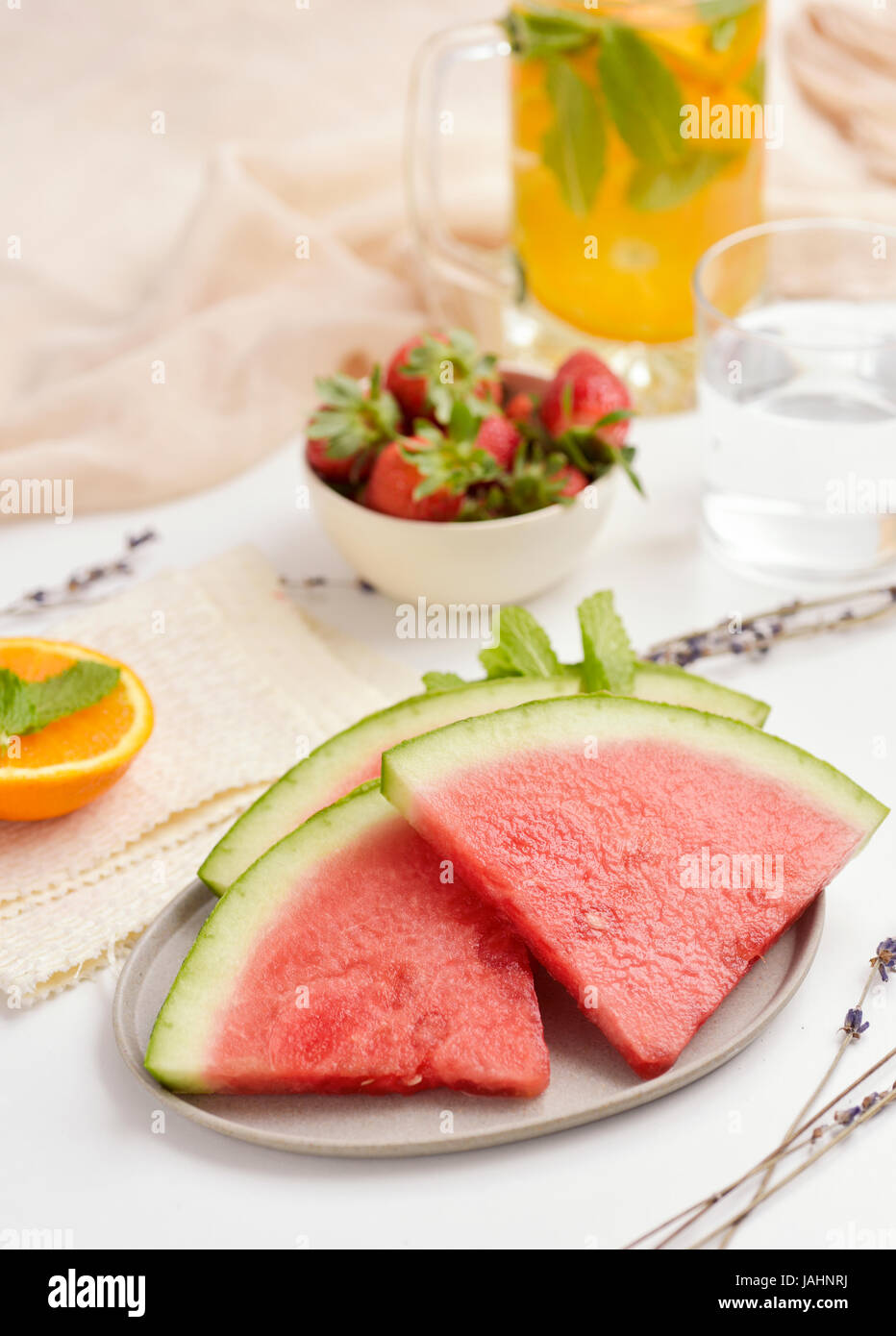 some slices of watermelon in a ceramic plate, a white ceramic bowl with strawberries and a jar with cold tea on a white a table Stock Photo