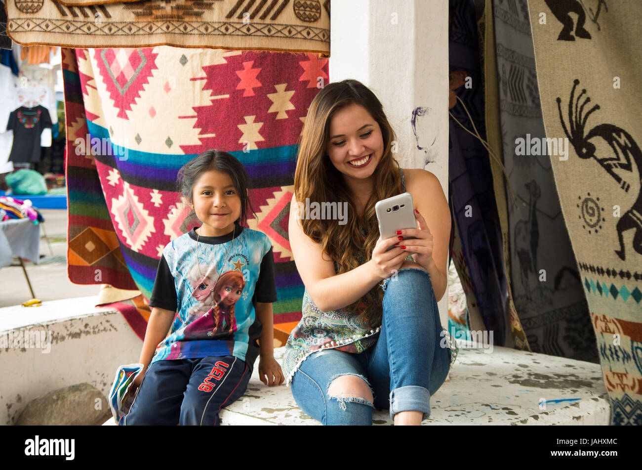 OTAVALO, ECUADOR - MAY 17, 2017: Beautiful young woman watching her cellphone next to an unidentified little indigenous girl, in colorful fabrics background Stock Photo