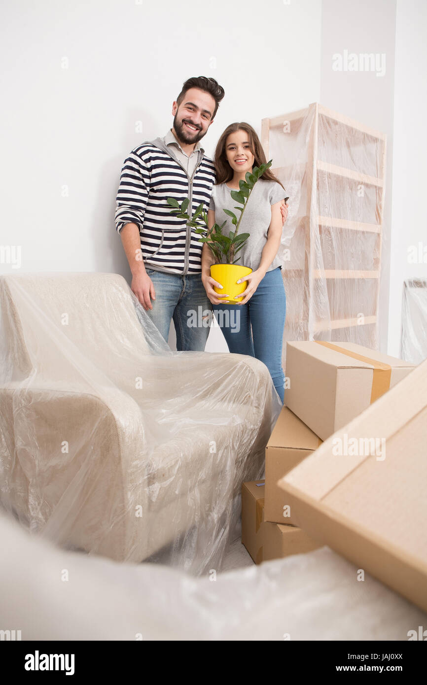 Relocating, yong couple standing in new apartment with furniture coverd with foil. Stock Photo