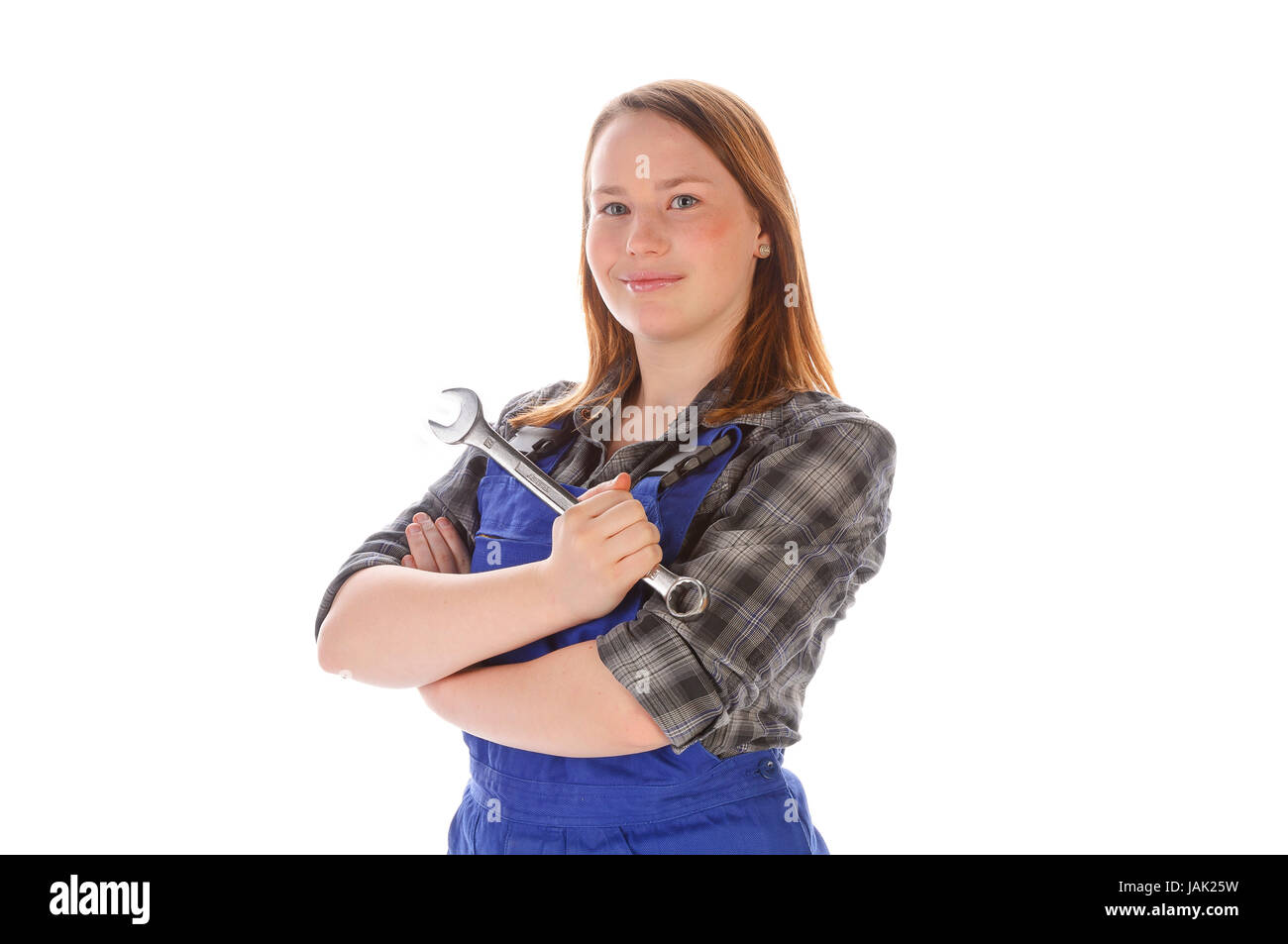 Junge Frau (Handwerkerin, Heimwerkerin, Auszubildende) mit langen Haaren trägt eine karierte Bluse und eine blaue Arbeits-Latzhose vor weißem Hintergrund. Sie hält einen großen Schraubenschlüssel (Maulschlüssel) in der Hand und lächelt zufrieden. Stock Photo