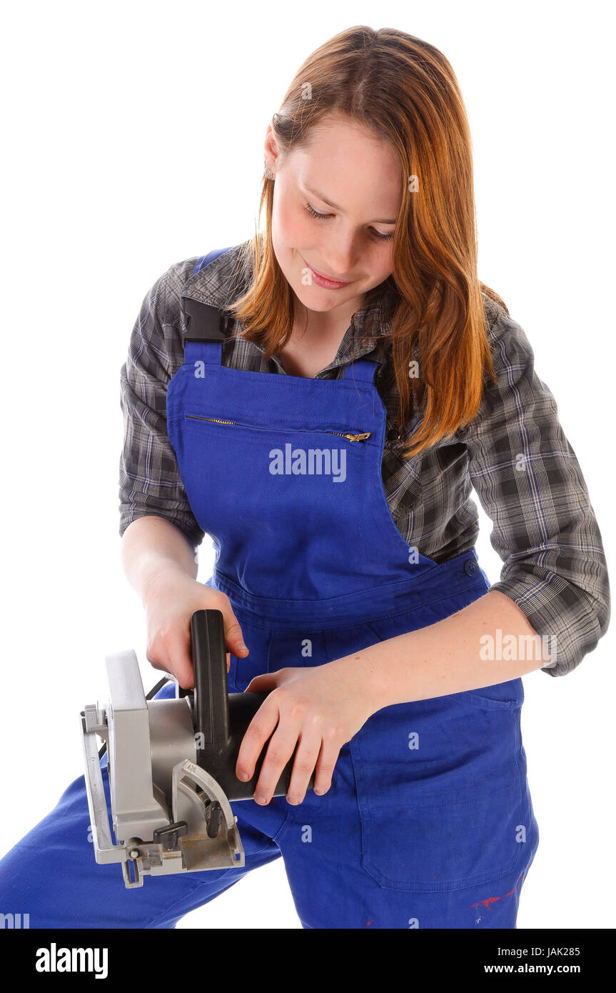 Junge Frau (Handwerkerin, Heimwerkerin, Auszubildende) mit langen Haaren trägt eine karierte Bluse und eine blaue Arbeits-Latzhose vor weißem Hintergrund. Sie hantiert mit einer elektrischen Kreissäge. Stock Photo