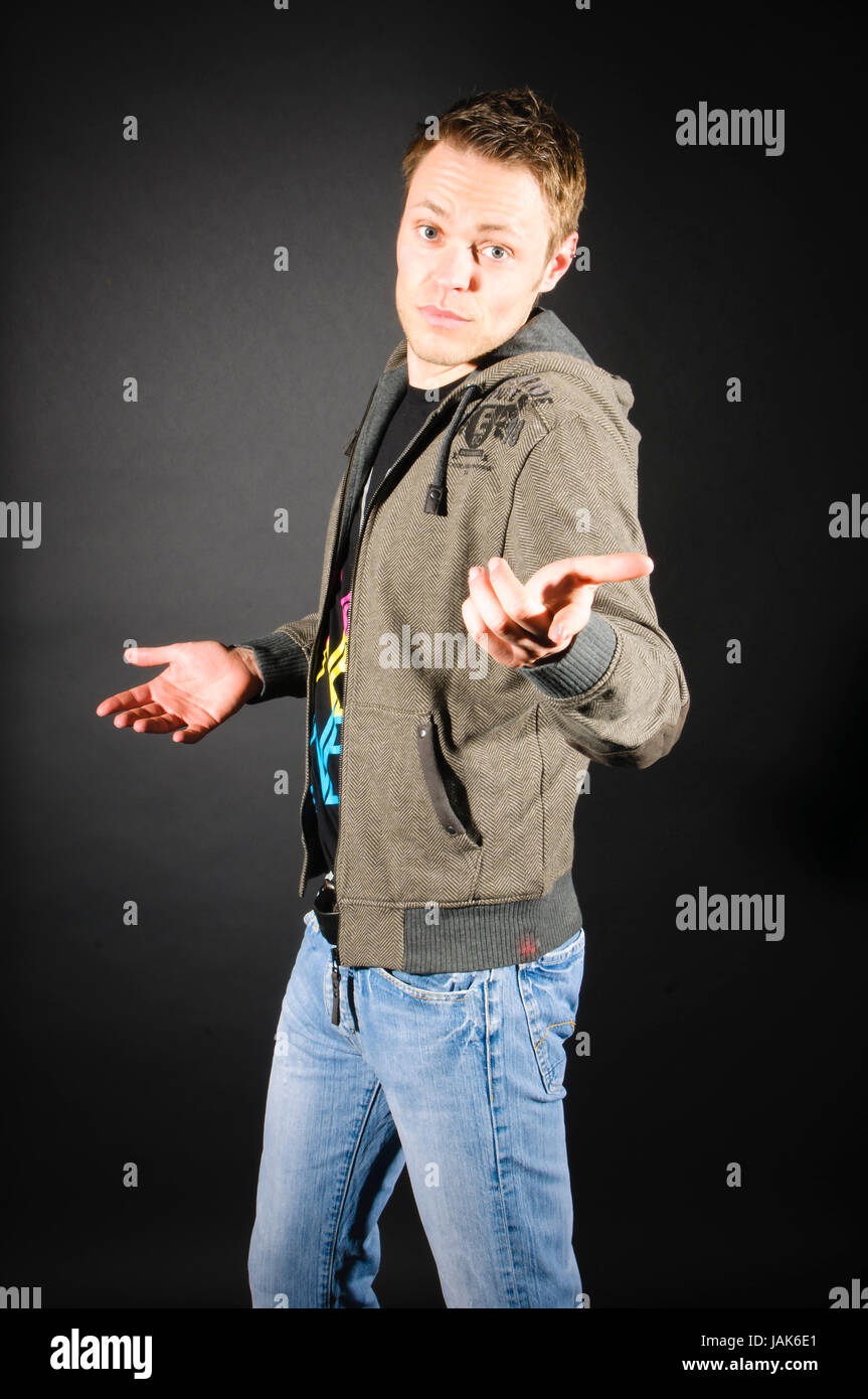 Junger Erwachsener Mann mit kurzen dunklen Haaren trägt Jeans und einen Kaputzenpullover und steht vor dunklem Hintergrund. Er macht eine unwissende Geste. Stock Photo