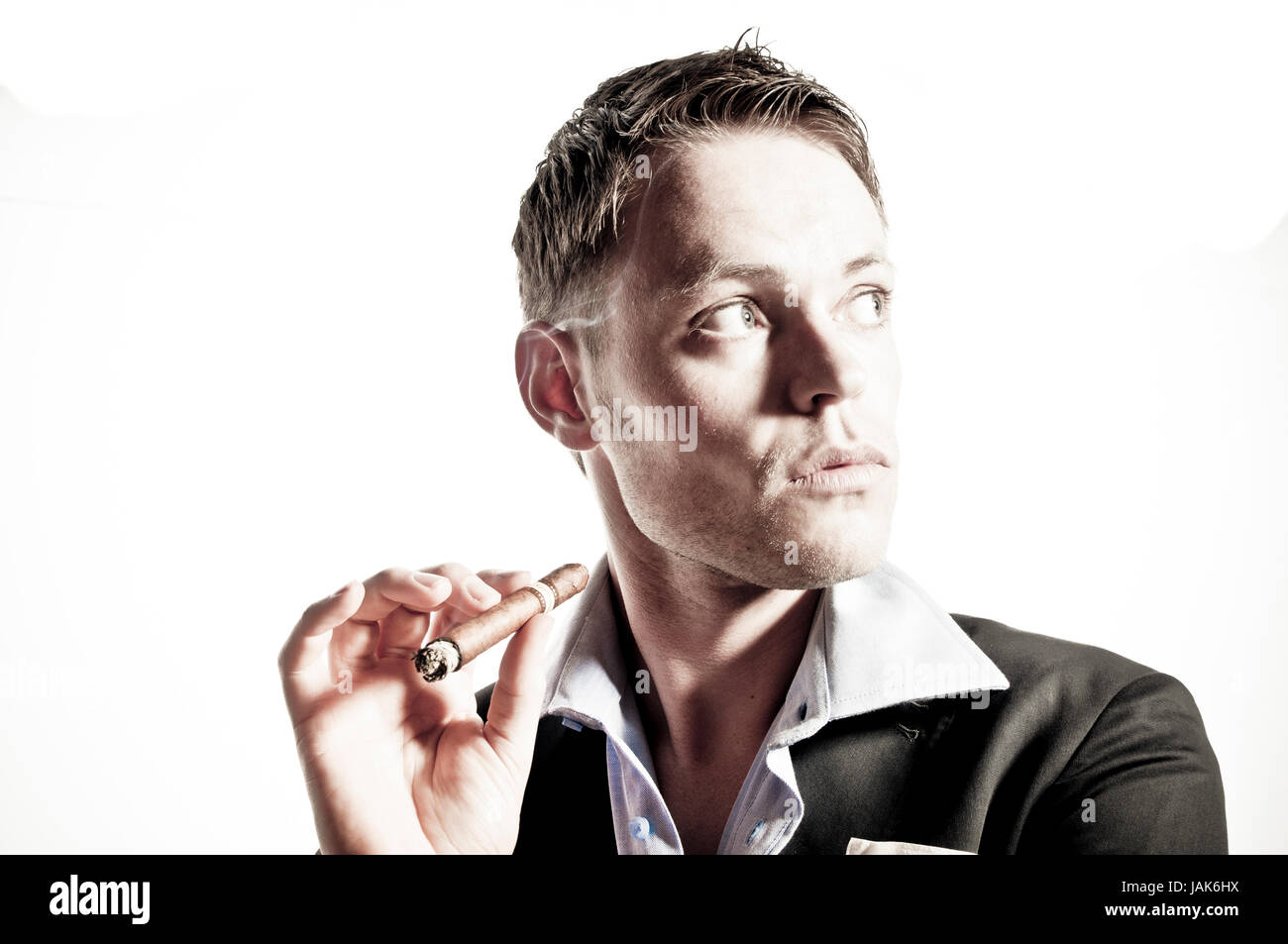 Junger Mann im dunklen Anzug raucht lässig eine Zigarre und schaut schräg in die Kamera vor weißem Hintergrund. Stock Photo