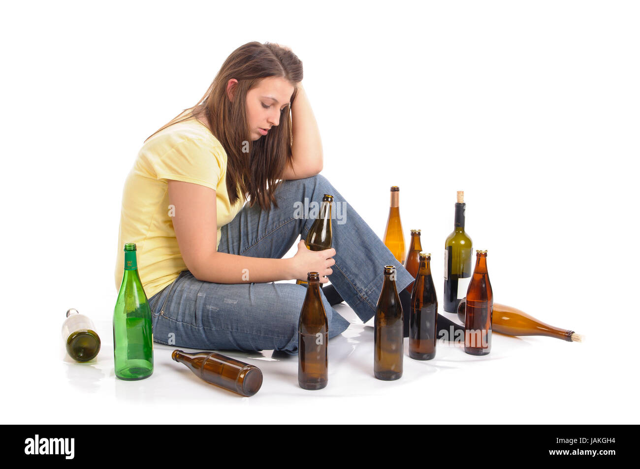 Junge Frau mit Jeans und T-Shirt sitzt auf dem Boden mit vielen leeren Bierflaschen und ist betrunken, isoliert vor weißem Hintergrund Stock Photo