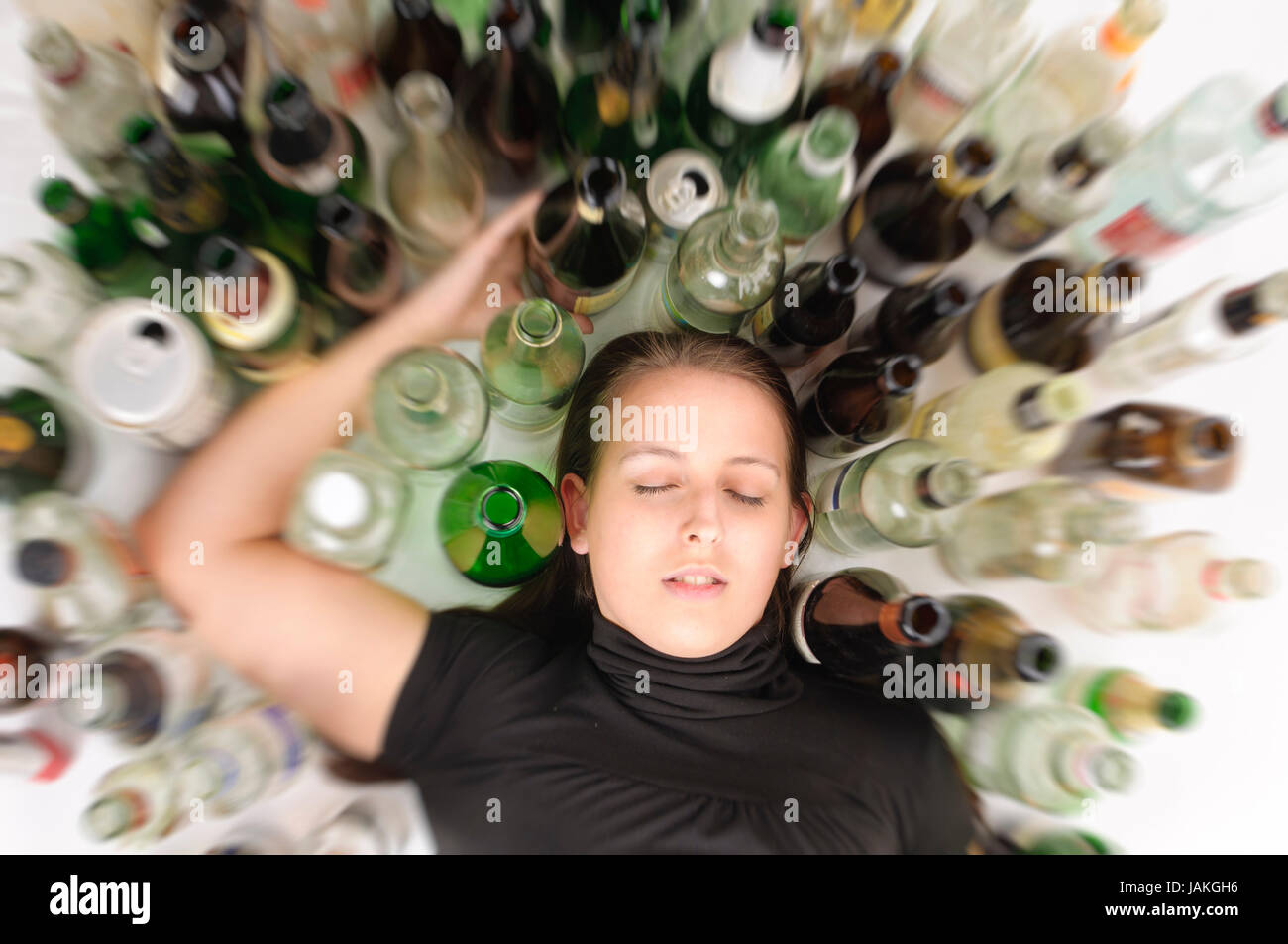 Junge Frau mit Jeans und T-Shirt sitzt auf dem Boden mit vielen leeren Bierflaschen und ist betrunken, fotografiert von oben, isoliert vor weißem Hintergrund Stock Photo