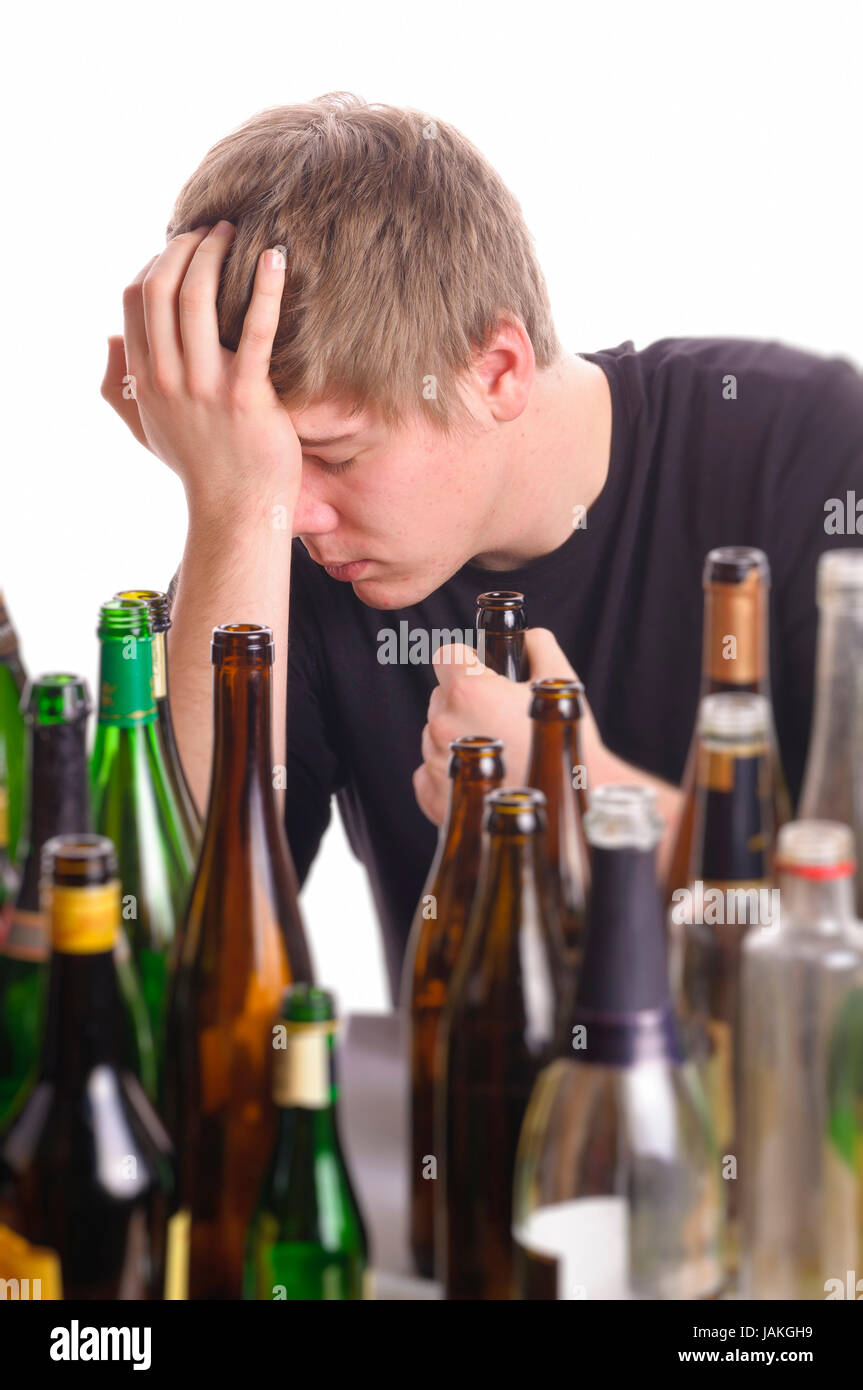 Junger Mann mit kurzen blonden Haaren sitzt am Tisch und ist von vielen leeren Bier- und Schnapfsflaschen umgeben, isoliert vor weißem Hintergrund. Stock Photo
