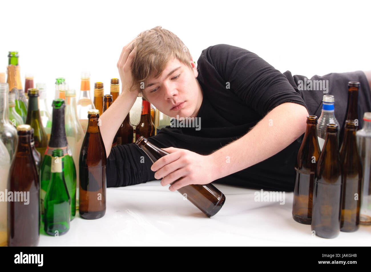 Junger Mann mit kurzen blonden Haaren liegt auf dem Boden und ist von vielen leeren Bier- und Schnapfsflaschen umgeben, isoliert vor weißem Hintergrund. Stock Photo