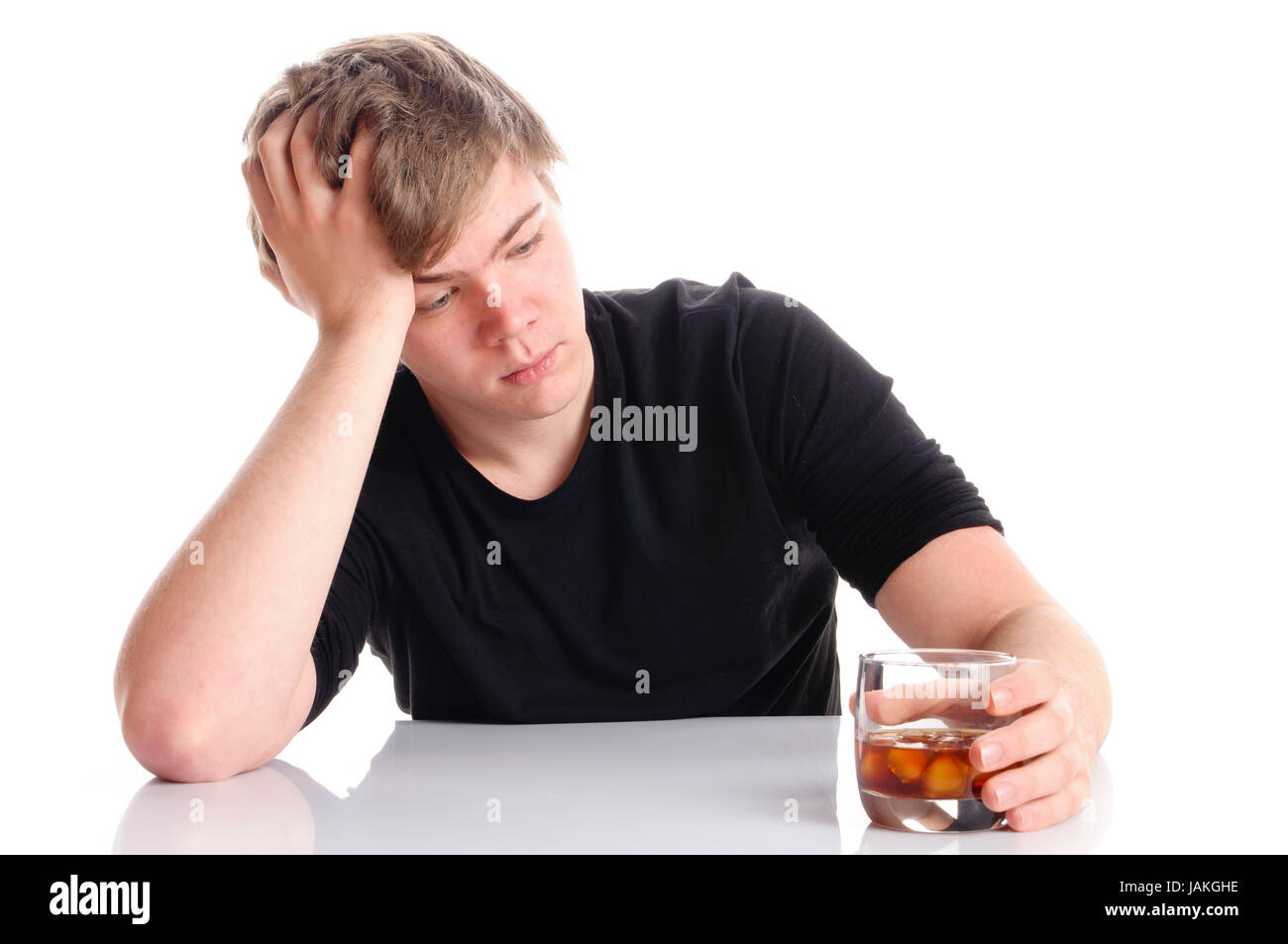 Junger Mann mit kurzen blonden Haaren und einem schwarzen T-Shirt sitzt an einem Tisch und hält Glas mit Branntwein in der Hand, isoliert vor weißem Hintergrund. Stock Photo