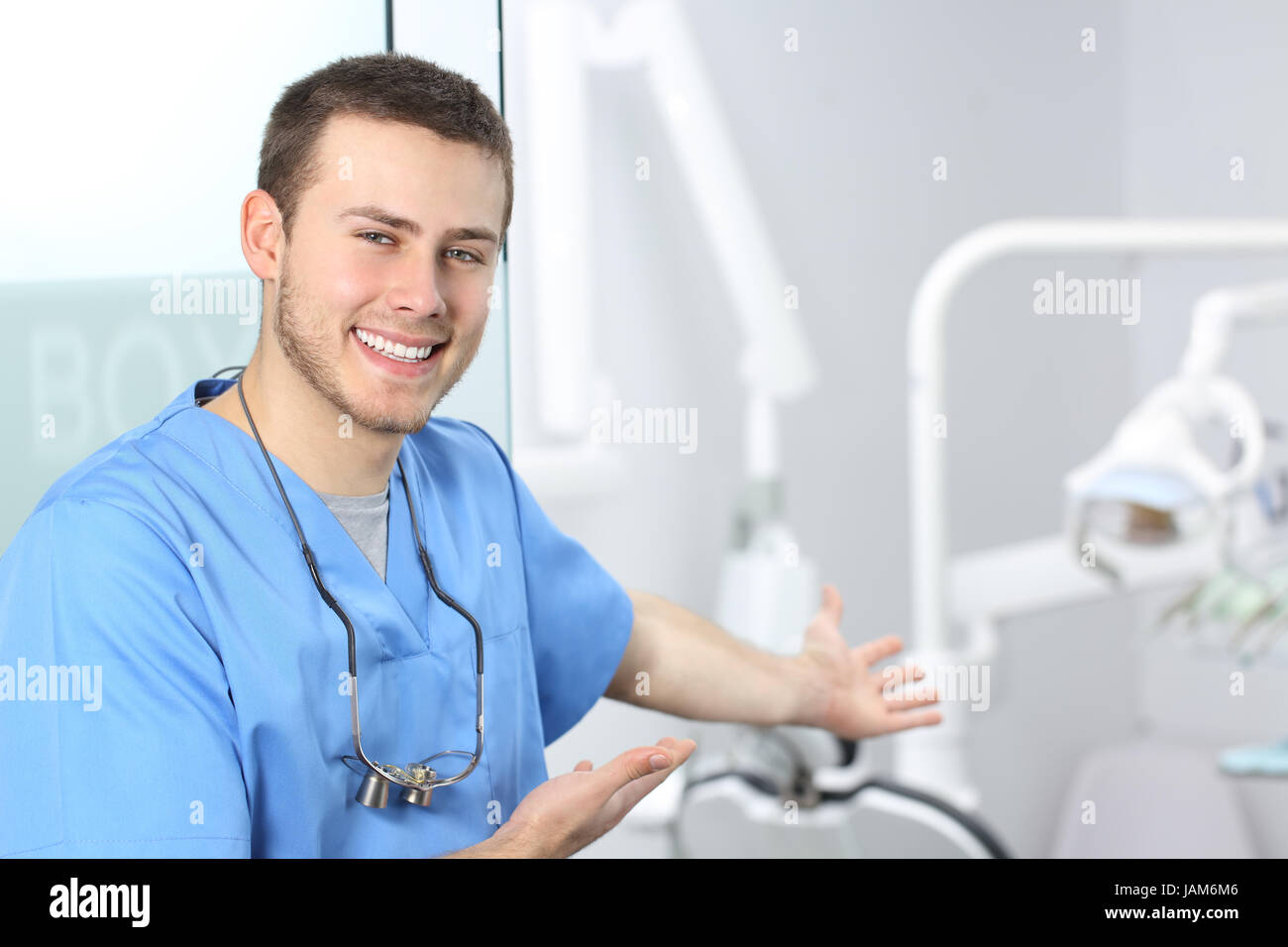 Doctor wearing blue coat inviting you to his dentist office and looking at camera with medical equipment in the background Stock Photo
