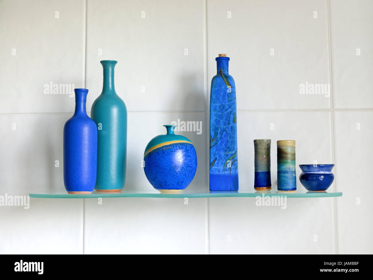 Still life arrangement of beautiful hand made artisan blue artisan pottery on a glass shelf, with a white tiled background. Stock Photo