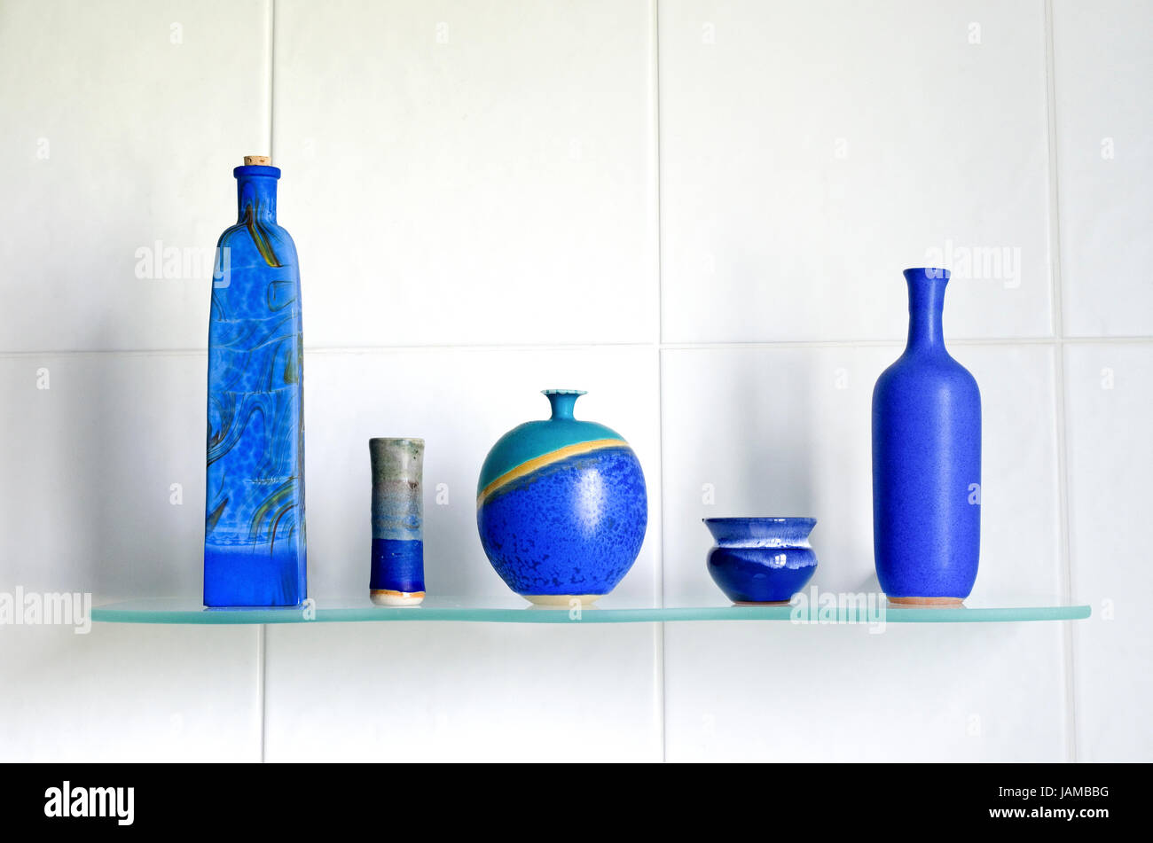 Still life arrangement of beautiful hand made artisan blue artisan pottery on a glass shelf, with a white tiled background. Stock Photo