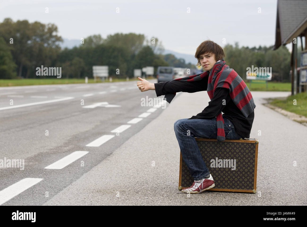 Roadside,teenager,boy,suitcase,sit,tear out autostopping,street,road marking,arrows,car,person,tread,at the side,jeans,pullovers,touched,Sneakers,luggage,travel,adventure,freedom,arm,hand,pollex,point,gesture,character,wait, Stock Photo