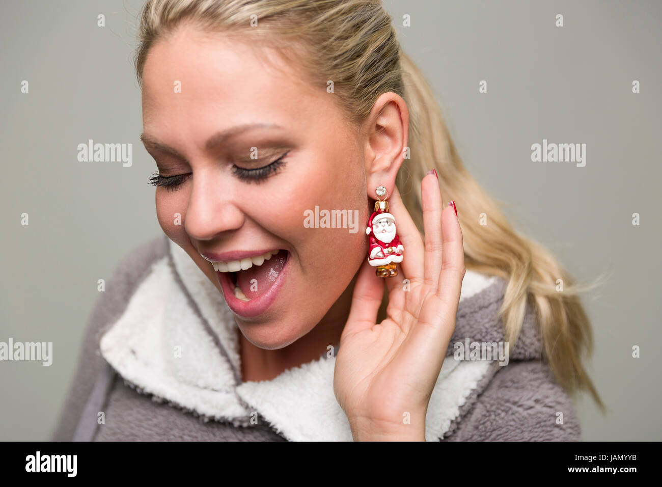 Frau mit blonden Haaren trägt Christbaumschmuck in Form eines Weihnachsmanss als Ohrring und zeigt diesen mit diesem die Vorfreude auf Weihnachten. Stock Photo