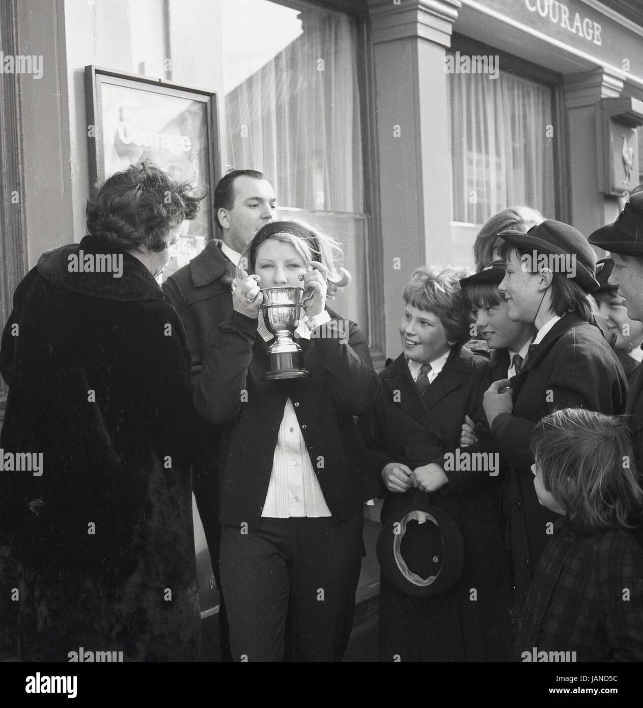 1965, historical, a young female shows off her trophy to the excited local school children, after winning the annual pancake race on the Old Kent Road, Peckham, London, between the Lord Wellington and Prince of Wales Pubs. Stock Photo