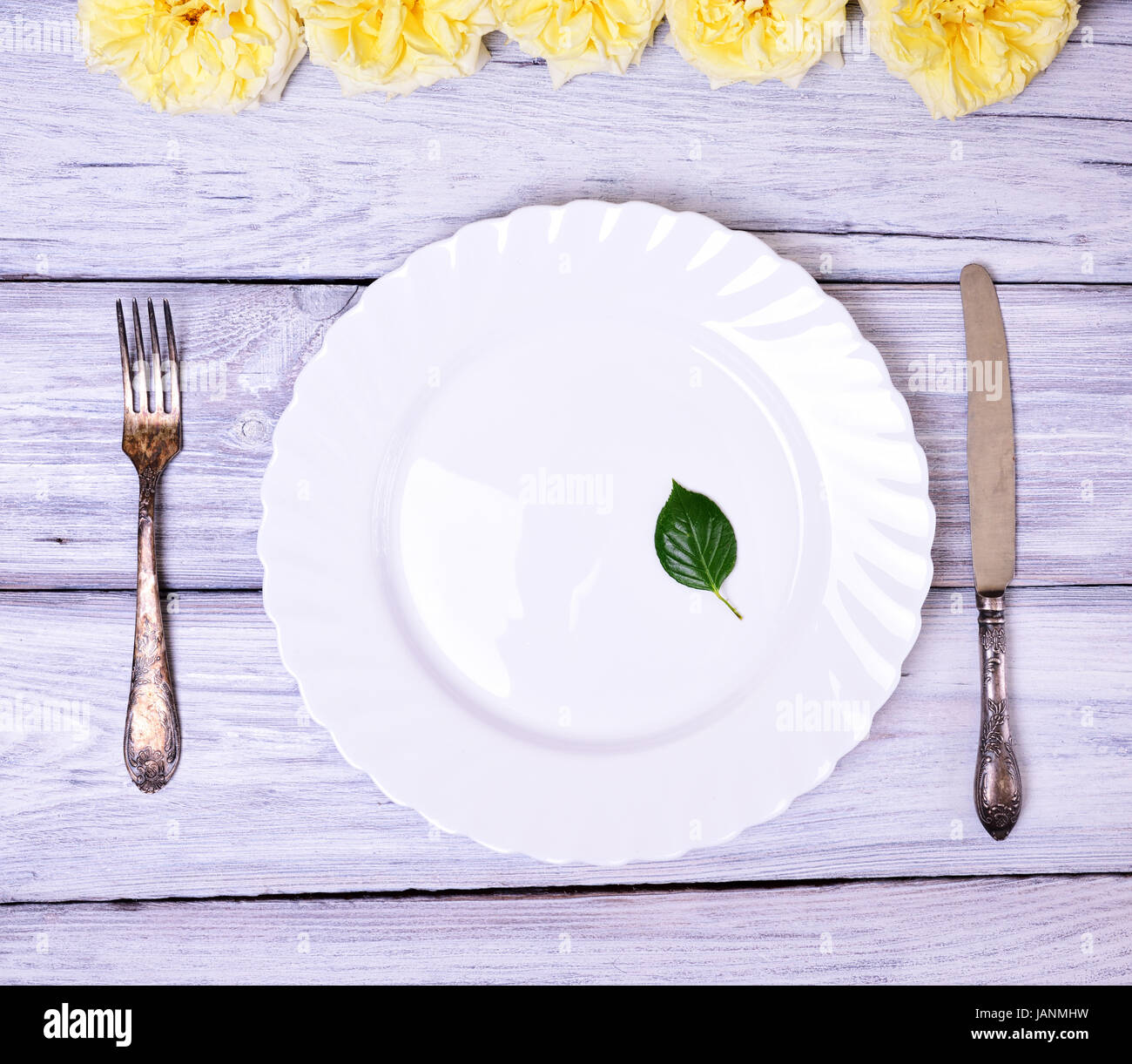 Empty white plate and metal fork and knife on a white wooden table, top