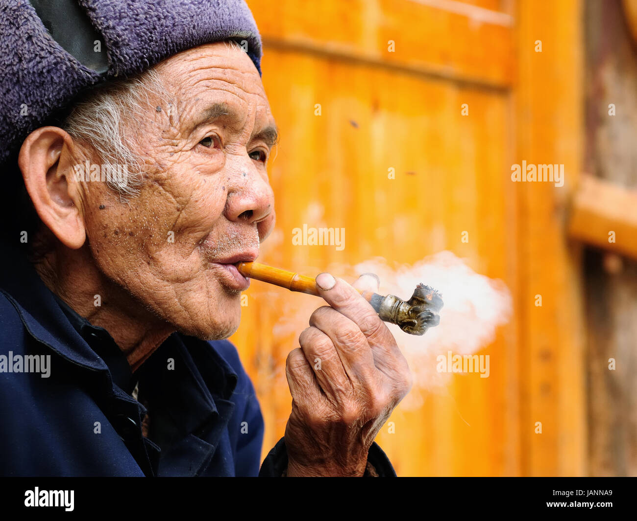 ZHAOXING, CHINA - 11 NOVEMBER 2010:  Old Chinese resting on the doorstep of his cottage smoking a pipe in the village Zhaoxing Stock Photo
