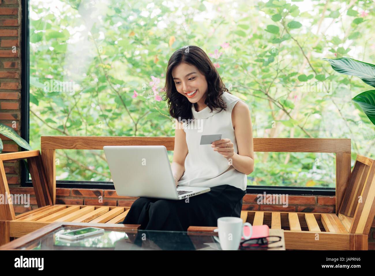 Confident young asian woman in smart casual wear shopping online while sitting near window in creative office or cafe Stock Photo