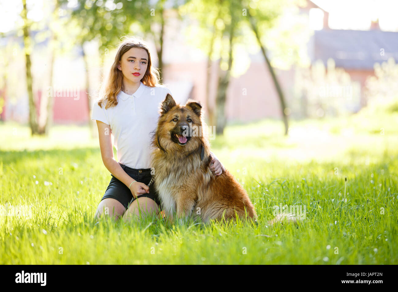 Teenage smiling girl in white shirt with her dog sitting on green grass ...