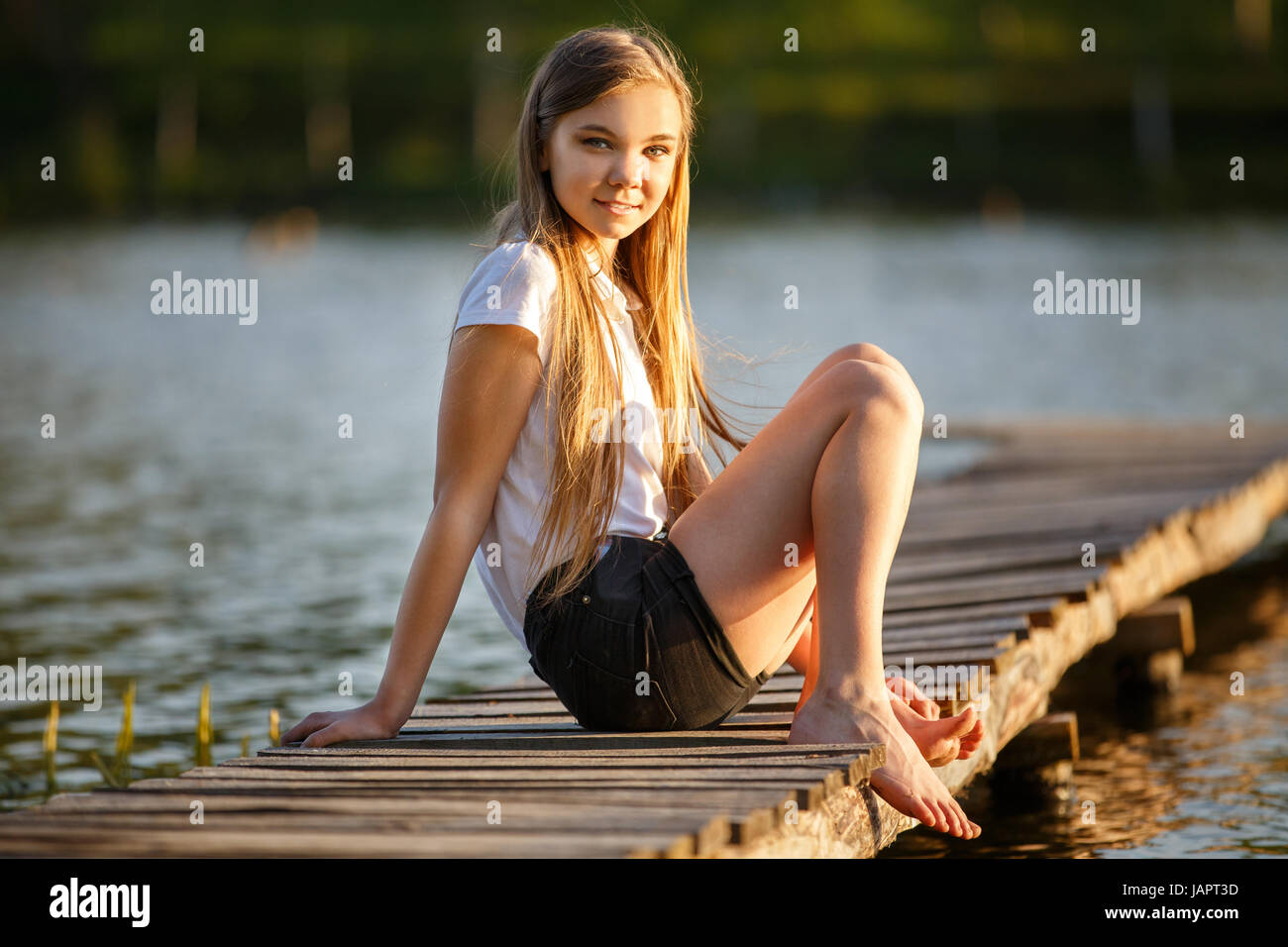 Young smiling girl sitting on pier near pond in sunset beams Stock Photo