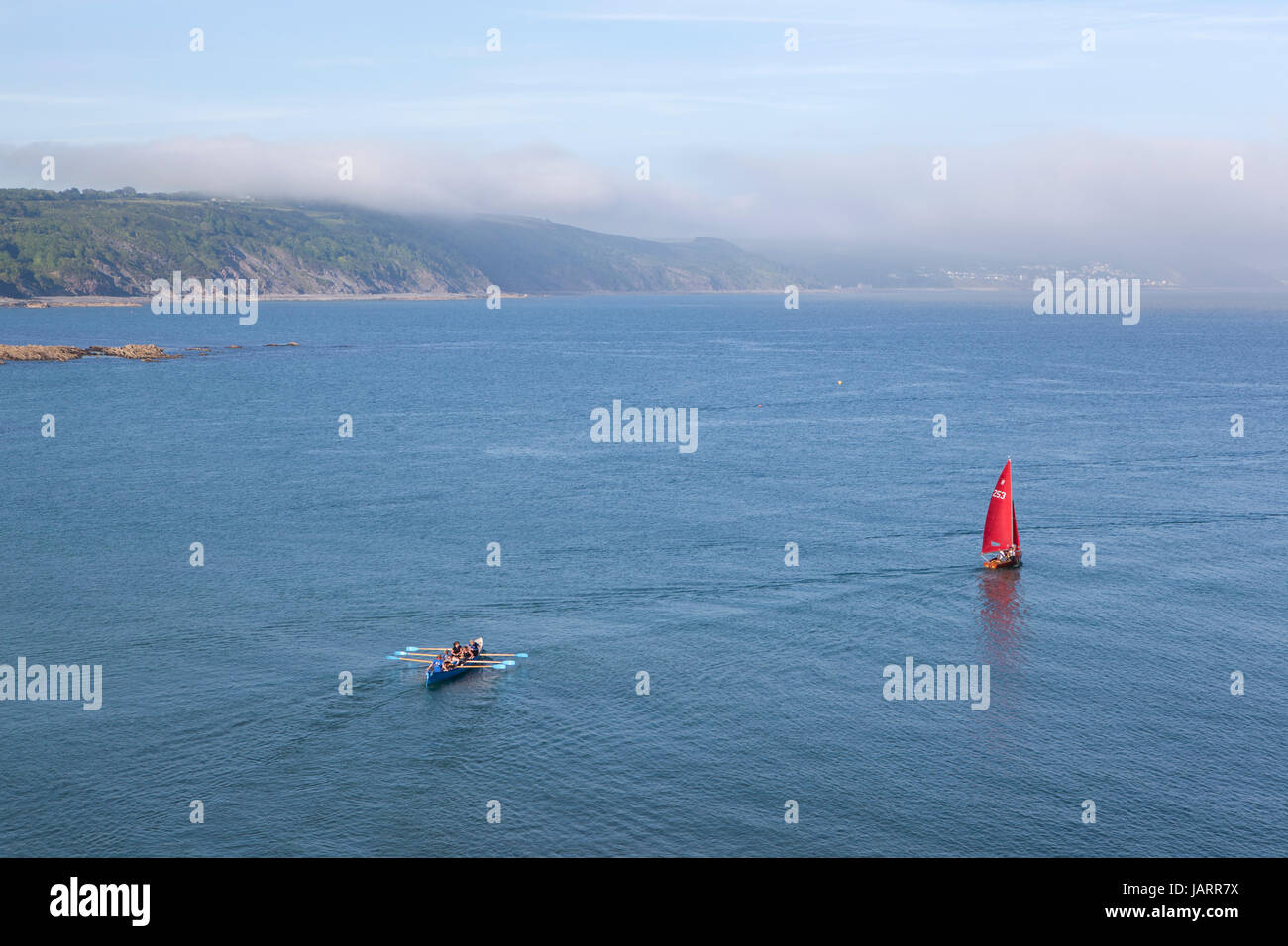 Sea mist rolls in to the coast at Looe, Cornwall as a red sailed dinghy and a pilot gig make their way out to sea. Stock Photo
