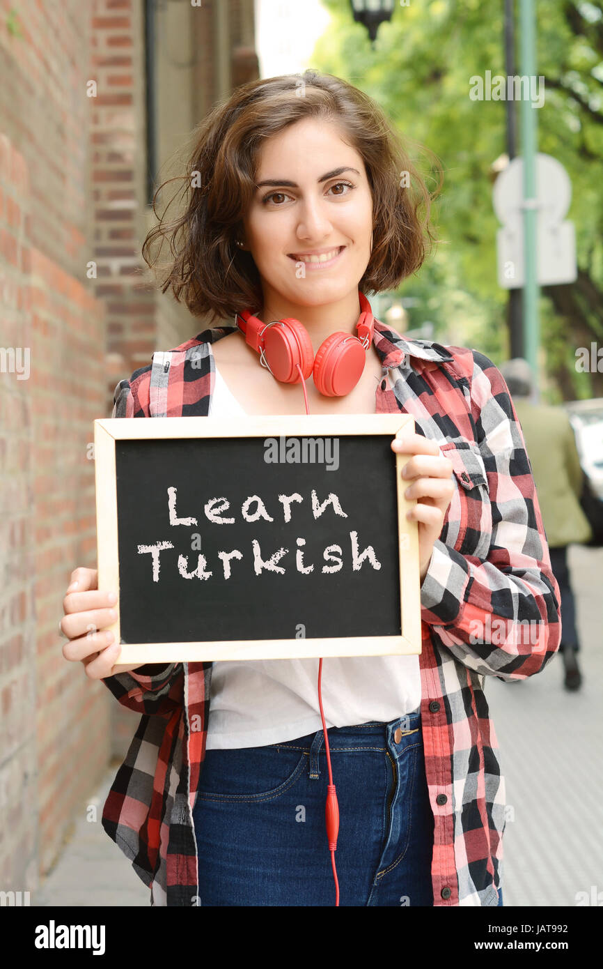 Young beautiful woman holding chalkboard with text 'learn turkish'. Education and learn foreign languages concept. Outdoors. Stock Photo
